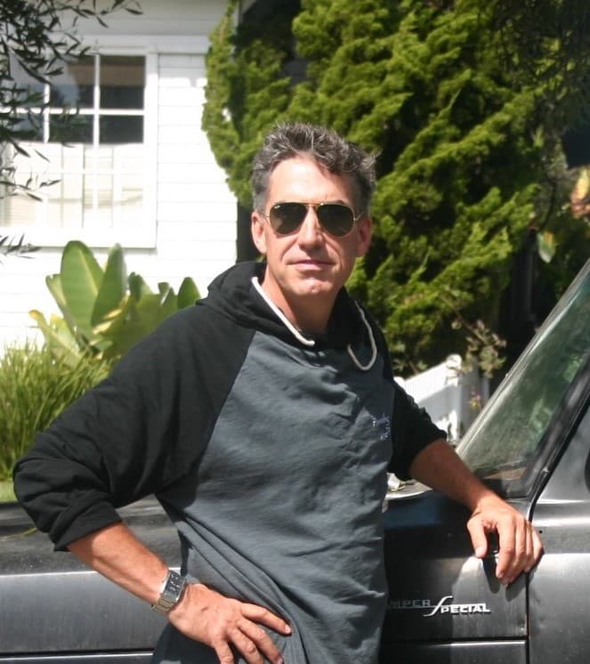 Bob Hayes leaning against a vintage truck in front of a craftsman home, in sunglasses and a hooded jacket.