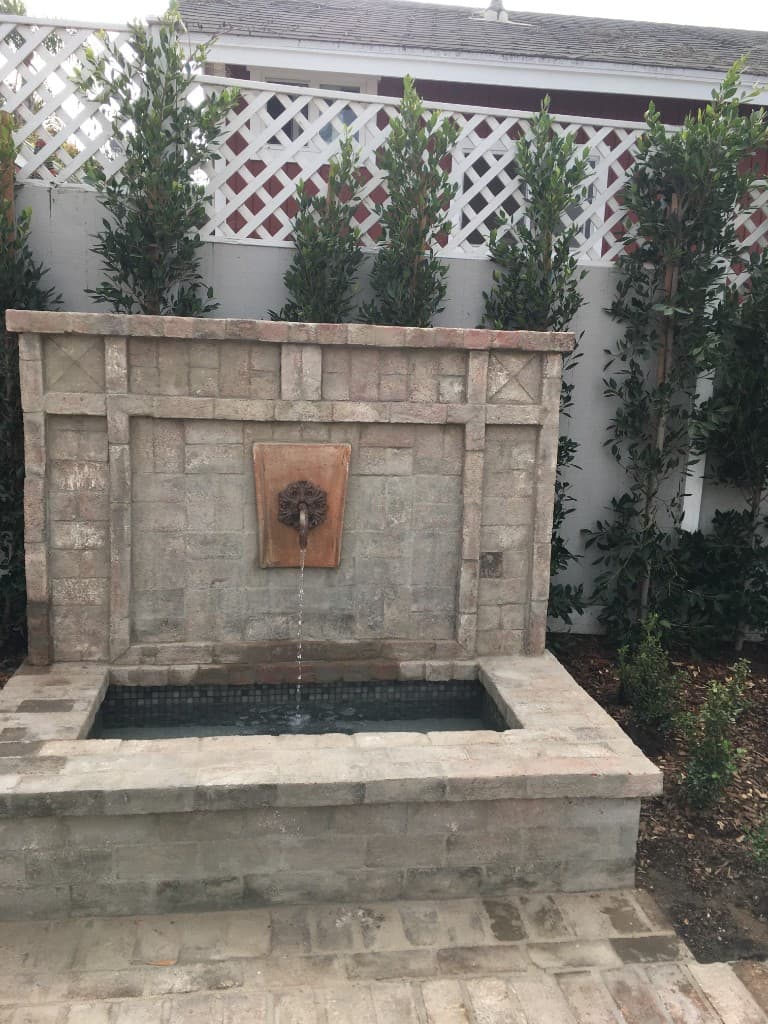 Custom stone wall fountain with a copper spout, framed by topiary, against a white lattice fence.